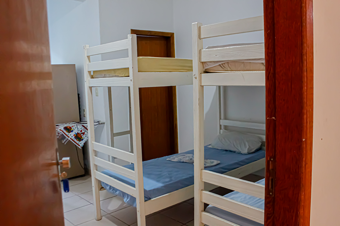 imagem mostra duas camas uma de casal e um beliche, as paredes brancas de um quarto, nas camas tem um lençol florido