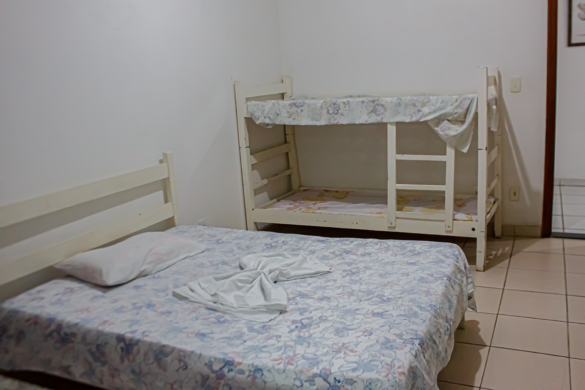 imagem mostra duas camas uma de casal e um beliche, as paredes brancas de um quarto, nas camas tem um lençol florido