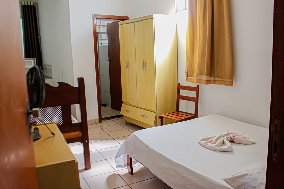 a imagem de um quarto com paredes brancas, onde aparece uma cama de casal, uma comoda branca, um ventilador, a porta para o banheiro e uma geladeira