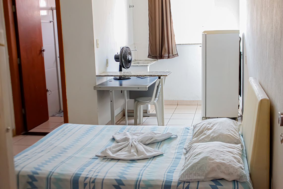 a imagem de um quarto com paredes brancas, onde aparece uma cama de casal, uma comoda branca, um ventilador, a porta para o banheiro e uma geladeira