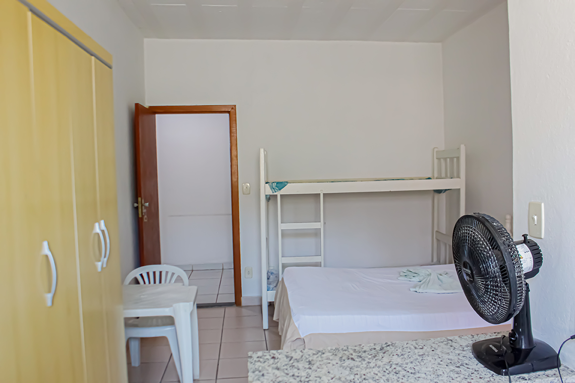 a imagem de um quarto com paredes brancas, onde aparece uma cama de casal, uma comoda branca, um ventilador, a porta para o banheiro e uma geladeira