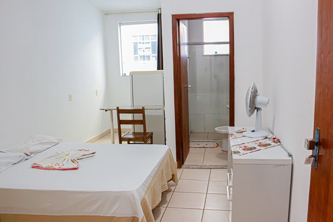 a imagem de um quarto com paredes brancas, onde aparece uma cama de casal, uma comoda branca, um ventilador, a porta para o banheiro e uma geladeira