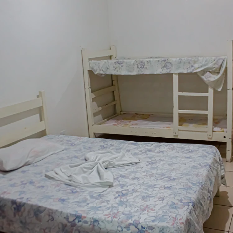 imagem mostra duas camas uma de casal e um beliche, as paredes brancas de um quarto, nas camas tem um lençol florido