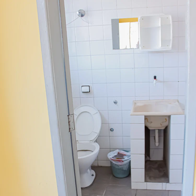 mostra um banheiro, onde é visivel um vaso sanitário na cor branca, um tapete de croche vermelho com branco, uma pia com bojo em inox, um box de vidro cristralino