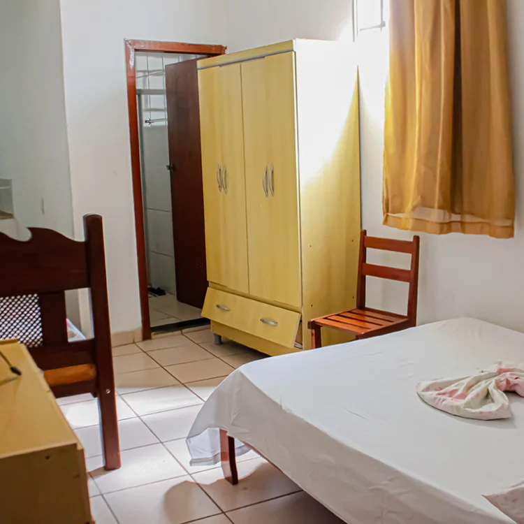 a imagem de um quarto com paredes brancas, onde aparece uma cama de casal, uma comoda branca, um ventilador, a porta para o banheiro e uma geladeira