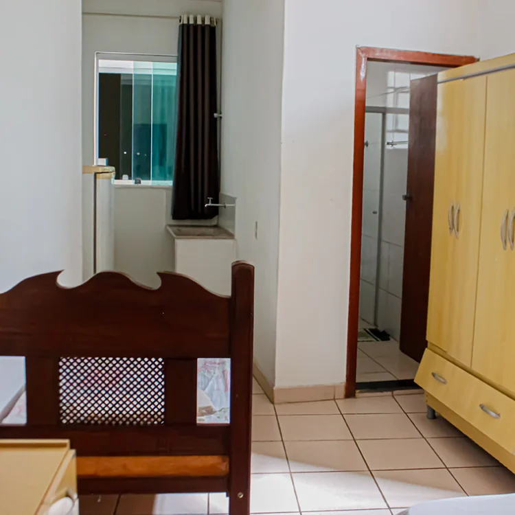 a imagem de um quarto com paredes brancas, onde aparece uma cama de casal, uma comoda branca, um ventilador, a porta para o banheiro e uma geladeira