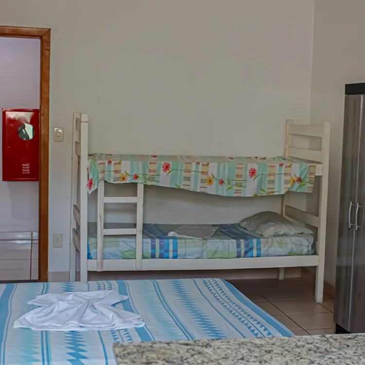 a imagem de um quarto com paredes brancas, onde aparece uma cama de casal, uma comoda branca, um ventilador, a porta para o banheiro e uma geladeira