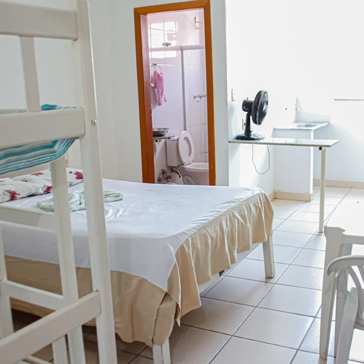 a imagem de um quarto com paredes brancas, onde aparece uma cama de casal, uma comoda branca, um ventilador, a porta para o banheiro e uma geladeira