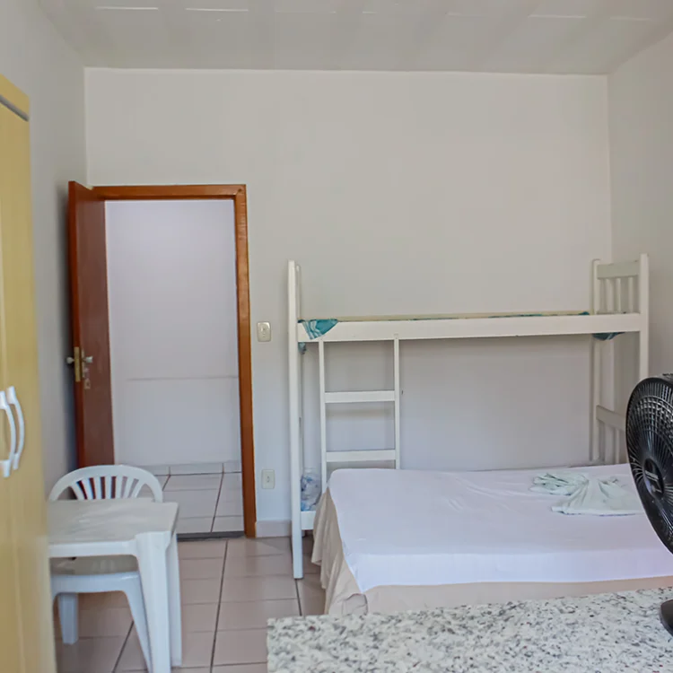 a imagem de um quarto com paredes brancas, onde aparece uma cama de casal, uma comoda branca, um ventilador, a porta para o banheiro e uma geladeira