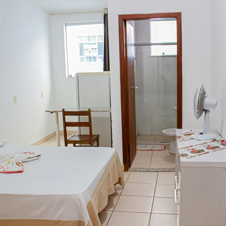 a imagem de um quarto com paredes brancas, onde aparece uma cama de casal, uma comoda branca, um ventilador, a porta para o banheiro e uma geladeira