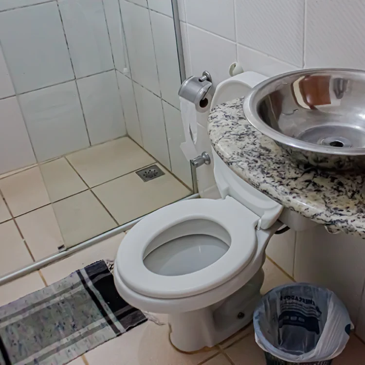 mostra um banheiro, onde é visivel um vaso sanitário na cor branca, um tapete de croche vermelho com branco, uma pia com bojo em inox, um box de vidro cristralino